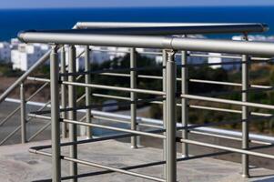 A Sleek Metal Railing Overlooking a Serene Coastal View With White Buildings Dotted Along the Horizon Near the Shore photo