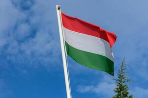 Hungarian flag waving proudly against a clear blue sky with a few clouds visible in the background during a bright sunny day photo
