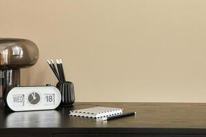 Stylish composition of office interior with black desk, table lamp, white clock, notebook and desk organizer. Template. Beige wall. Copy space. photo