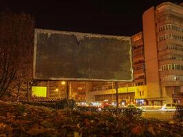 Empty billboard in urban area at night surrounded by illuminated buildings and fall leaves. photo