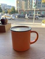 Orange stylish ceramic cup of cappuccino or latte decorated with foam on overlooking the street and the road and moving cars. Morning coffee. Warm Coffee Moment in a Cozy Caf Setting photo