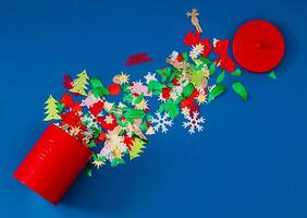 Colorful Christmas glitter confetti mix falling from red Xmas cookies tin decorated with classic bells on blue background. Minimal Christmas and New Year concept. Creative festive holiday flat lay. photo