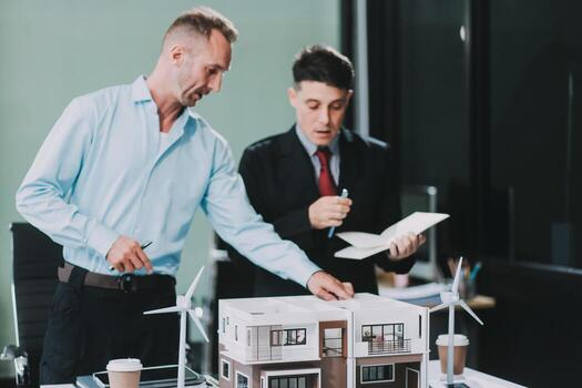 Engineers meeting about wind turbine model and house model. Renewable energy and housing development concept. Team of architects and engineers in modern office discussing design structure. photo