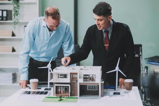 Engineers meeting about wind turbine model and house model. Renewable energy and housing development concept. Team of architects and engineers in modern office discussing design structure. photo
