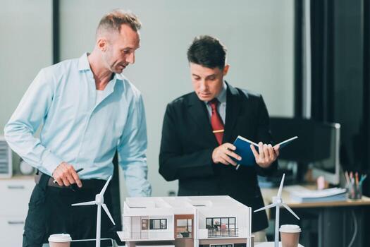 Engineers meeting about wind turbine model and house model. Renewable energy and housing development concept. Team of architects and engineers in modern office discussing design structure. photo