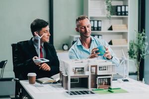 Engineers meeting about wind turbine model and house model. Renewable energy and housing development concept. Team of architects and engineers in modern office discussing design structure. photo