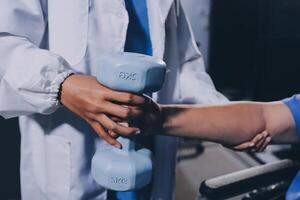 Old woman training with physiotherapist using dumbbells at home. Therapist assisting senior woman with exercises in nursing home. Elderly patient using dumbbells with outstretched arms. photo