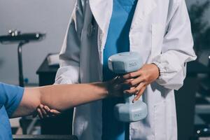 Old woman training with physiotherapist using dumbbells at home. Therapist assisting senior woman with exercises in nursing home. Elderly patient using dumbbells with outstretched arms. photo