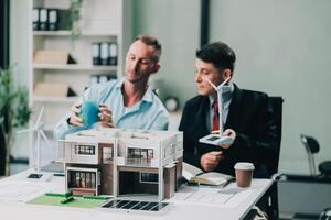 Engineers meeting about wind turbine model and house model. Renewable energy and housing development concept. Team of architects and engineers in modern office discussing design structure. photo
