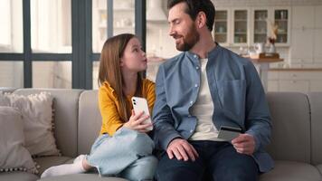 Joyful family little Caucasian girl daughter choose goods online shop with mobile phone dad man parent hold credit card clients kid father use bank app for successful payment male hug child smile home video