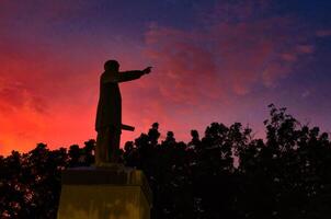 A statue of a man holding a gun in front of a colorful sunset photo