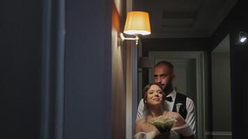 Romantic Couple Embracing in Dim Hallway, A newlywed couple stands close in a dimly lit hallway, the groom embracing the bride from behind as she holds a bouquet video