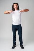 A young man with curly hair joyfully pointing down while wearing a plain white t-shirt against a neutral background photo