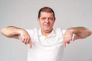 A man in a white shirt smiling and gesturing downwards in a studio with a light gray background photo
