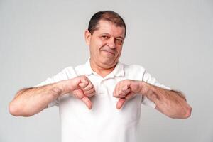 Older man expressing disapproval with thumbs down gesture while standing against a plain background photo