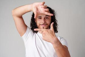 A young man with curly hair framing his face and making a gesture as if capturing a moment against a plain background photo