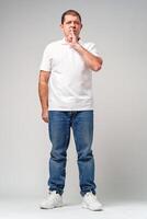 A man makes a playful gesture with his hand near his mouth while smiling in a neutral backdrop photo