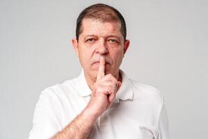 A man makes a playful gesture with his hand near his mouth while smiling in a neutral backdrop photo