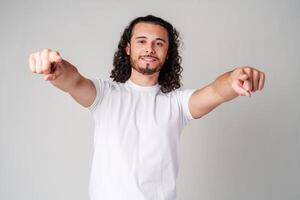 A smiling young man with curly hair playfully pointing directly at the viewer in a light-colored setting photo