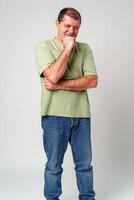 A man in a light green shirt and jeans stands against a plain background with a thoughtful expression photo
