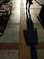 A person is walking on a sidewalk next to a bench. The bench is empty. The sidewalk is lined with bricks and has a shadow cast by the person photo
