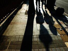 A person is walking on a sidewalk next to a bench. The bench is empty. The sidewalk is lined with bricks and has a shadow cast by the person photo