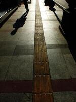 A person is walking on a sidewalk next to a bench. The bench is empty. The sidewalk is lined with bricks and has a shadow cast by the person photo