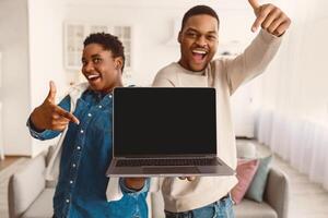 Check This Website. Excited African American couple holding and showing laptop with black empty screen in hands, presenting and pointing at gadget with blank space for mockup, blurred background photo