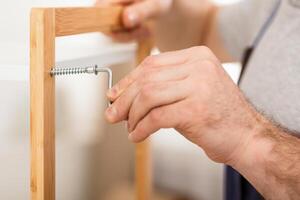 Closeup Of Unrecognizable Carpenter's Hands While He Tightening Screws Working Assembling Shelf Indoors, Wearing Coverall Uniform. Carpentry Service Concept. Cropped Shot photo