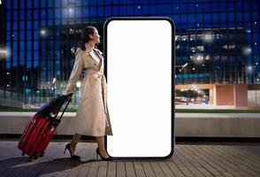 A woman carrying a red suitcase strolls by a massive blank smartphone screen in an urban setting during evening hours. The lights from nearby buildings create a vibrant atmosphere. photo