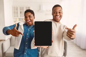 Great Application. Excited African American couple holding and showing digital tablet with black empty screen in hands, presenting and pointing at gadget with blank space for mockup, selective focus photo