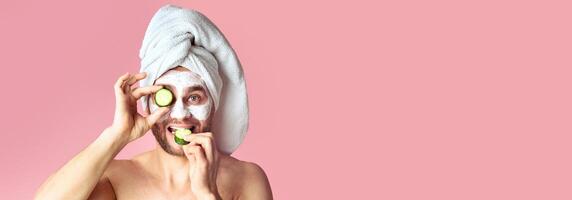 Millennial man makes face mask and eats cucumber on pink background photo