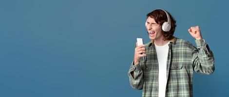 Trendy teenager in headset using smartphone as mike, singing along with his favorite song, color background. Copy space photo