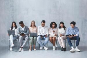 Multiracial young group of people sitting in line and waiting for job interview, talking to each other and looking at laptops, panorama photo