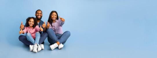Smiling African American family of three showing thumbs up gesture with both hands, approving or recommending something good, sitting on the floor at blue studio background, panorama, free copy space photo