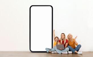 A mother, grandmother and granddaughter are seated on the floor, cheerfully posing beside a giant blank smartphone. They are engaged in playful interaction and smiling at the camera. photo