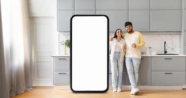 A cheerful Indian couple poses in a sleek kitchen, smiling while standing beside a large, blank smartphone display. They enjoy each other's company in a stylish home environment. photo