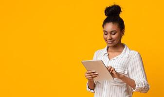 Modern Technologies For Life. Smiling African Girl Using Digital Tablet While Standing Over Yellow Background In Studio, Free Space photo