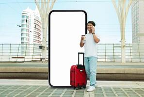 Asian young guy traveler stands with a red suitcase and a coffee cup by huge phone with blank screen, talking on the phone at a modern train station photo