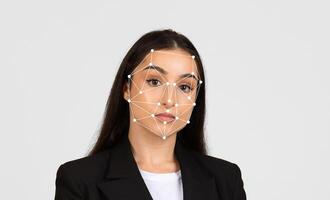 A woman with long black hair poses against a plain background while a facial recognition grid overlays her face. She appears professional, wearing a blazer and white top. photo