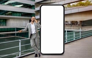 A businessman dressed in a gray suit is casually talking on his smartphone while leaning against a railing, standing by huge phone with blank screen photo
