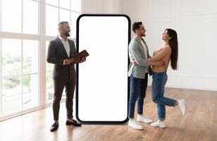 A couple shares a joyful moment in a bright, modern interior. They embrace while a man with a tablet observes, huge phone with blank screen next to them photo