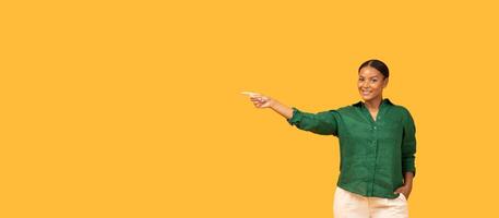 A black woman smiles and points to copy space while dressed in a green shirt and light-colored pants. The backdrop is a vibrant yellow, creating a lively and inviting atmosphere. photo