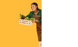 A young woman in an apron joyfully presents a wooden crate filled with fresh vegetables. The vibrant background enhances her cheerful demeanor while showcasing the bounty of fresh produce. photo