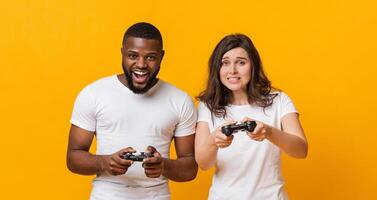 Gambling multiracial couple playing videogames with joysticks, posing together over yellow background in studio, guy winning, free space photo