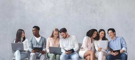 Group of multiracial young people sitting in line and waiting for job interview, talking to each other and looking at laptops, panorama photo