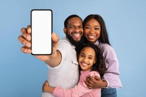 Trendy Mobile Phone. Portrait of happy family of three people of color holding smartphone with empty screen in hand, showing device with blank space for mockup close to camera, blue studio background photo