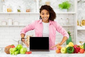 Website Template. Smiling african woman showing computer with blank monitor in loft kitchen, mock up, empty space photo