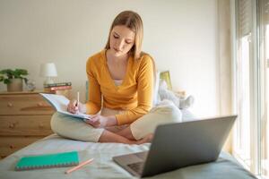 A teen girl sits cross-legged on her bed, focused on writing notes in a notebook. A laptop rests nearby on the bed, and sunlight filters through the room, creating a warm atmosphere. photo