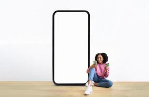 A black woman sits on the floor with a smile, sipping from two drinks, next to a large blank smartphone display. She wears a casual pink sweater and jeans, creating a relaxed atmosphere. photo
